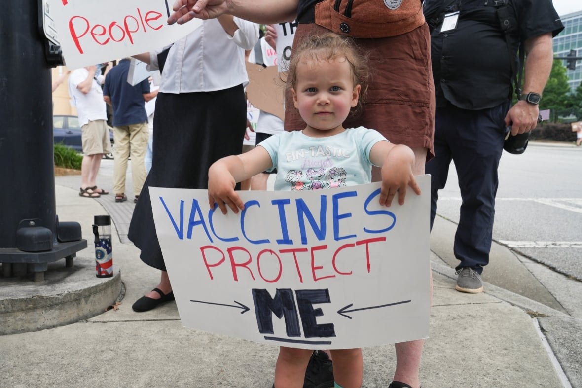 a young girl holds up a white placard that reads "Vaccines protect me". behind her a crowd of protesters gathers