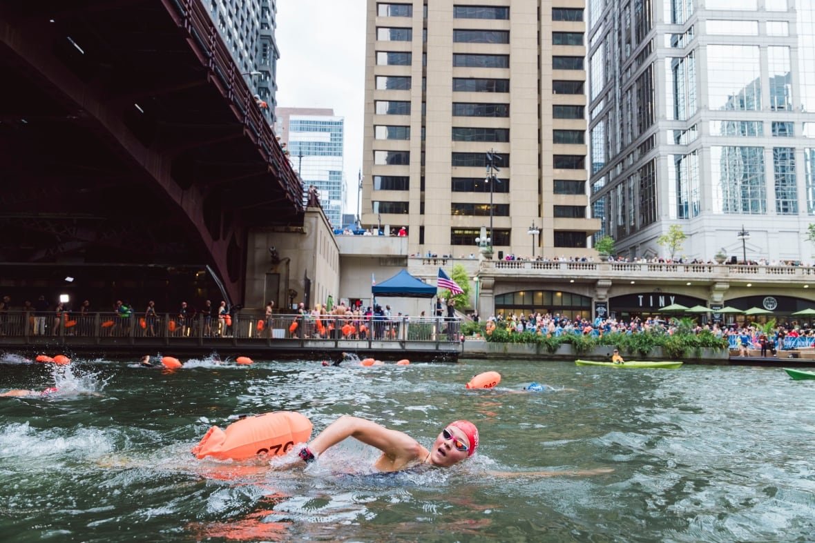 A woman in a swim cap and goggles does the front-crawl down a city river as people watch from a pedestrian walkway. 