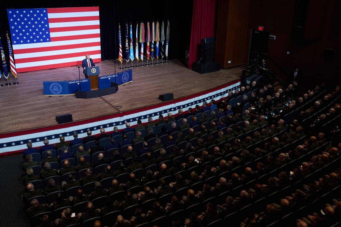 A man in a suit speaks from a podium in front of hundreds of military personnel seated in a theatre.
