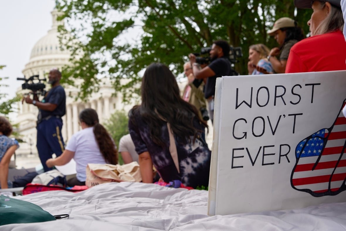 A white placard reading "worst government ever" sits on a white blanket on the ground in the foreground of the photo, in front of a crowd of people and a man speaking at a podium in front of a large white building with a tall dome on top.