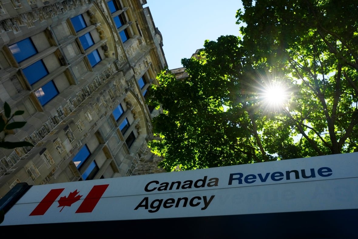 Sunlight shines through the green leaves of a tree in front of an older stone building. A sign in front reads "Canada Revenue Agency."