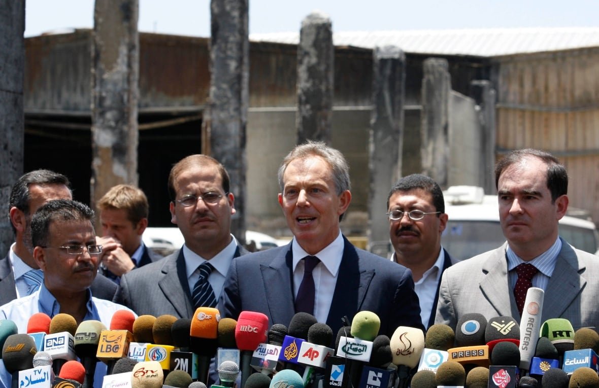 A man with short, grey hair wearing a dark suit is flanked by several other men in suits as he speaks in front of a grouping of news microphones.