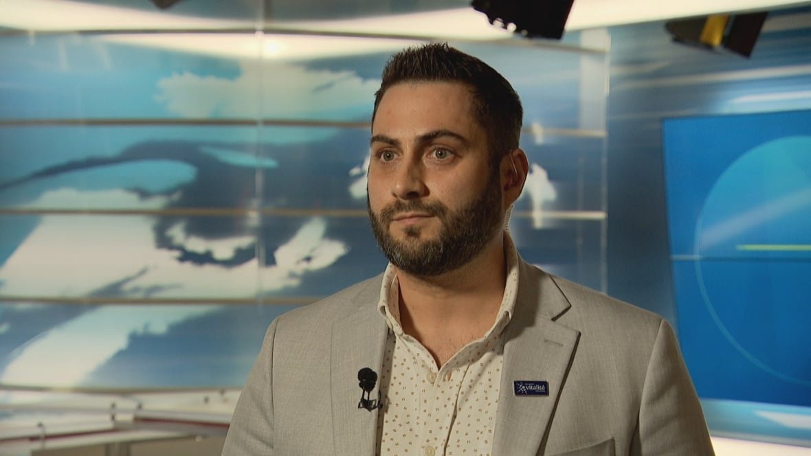 A man with dark hair, a beard and mustache, wearing a gray suit with a white collared shirt, looks to the side while standing in a TV studio.