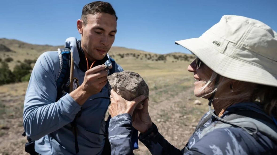 A man with short brown hair and a blue jacket looks intently at a rock that a woman is holding up in front of him in a desert region.