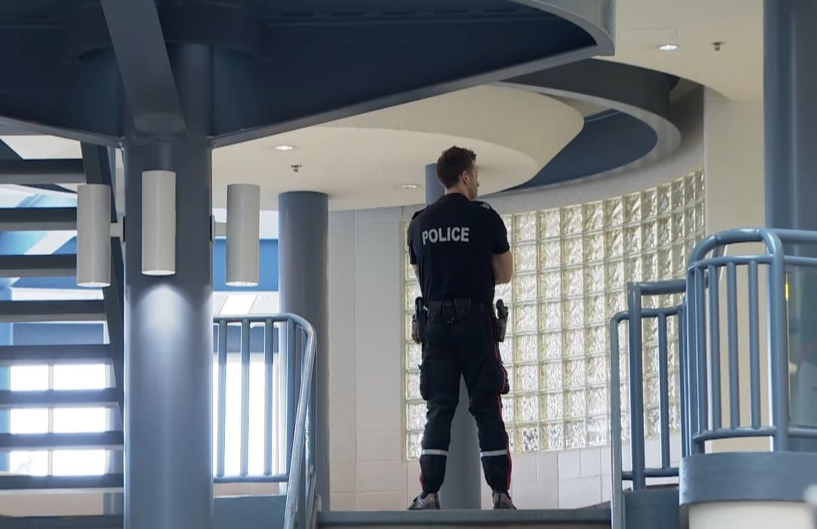 A man in a police uniform stands with his back to the camera at the top of a flight of stairs in a hallway next to some windows. 