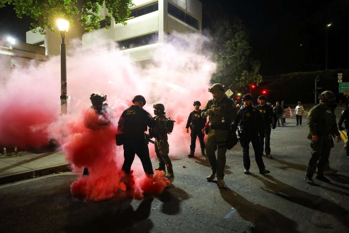 Red smoke billows around people on the street at night in front of a large white building. Some wear military uniforms.