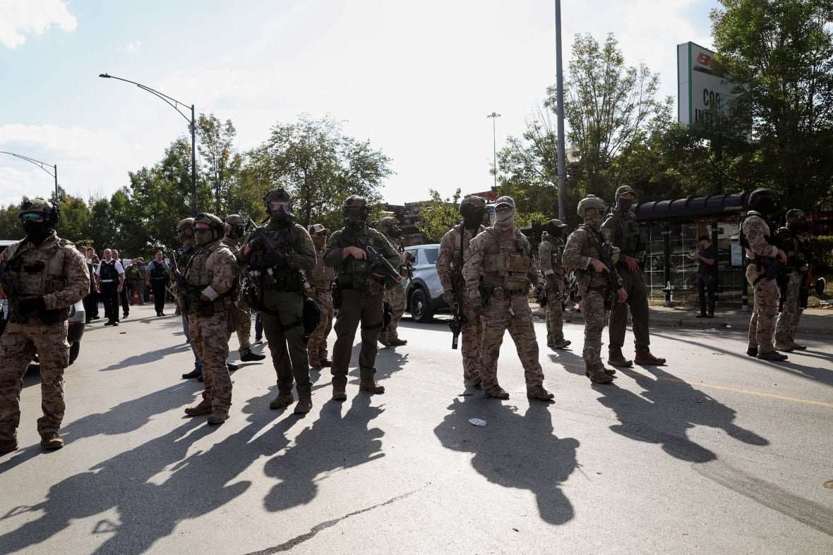 Masked men in military uniforms, holding guns, stand on the street in the bright sun.