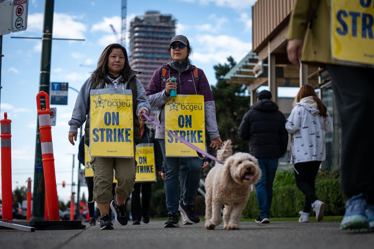 Two women wearing BCGEU strike placards, one walking a small white dog, picket outside a liquor strore