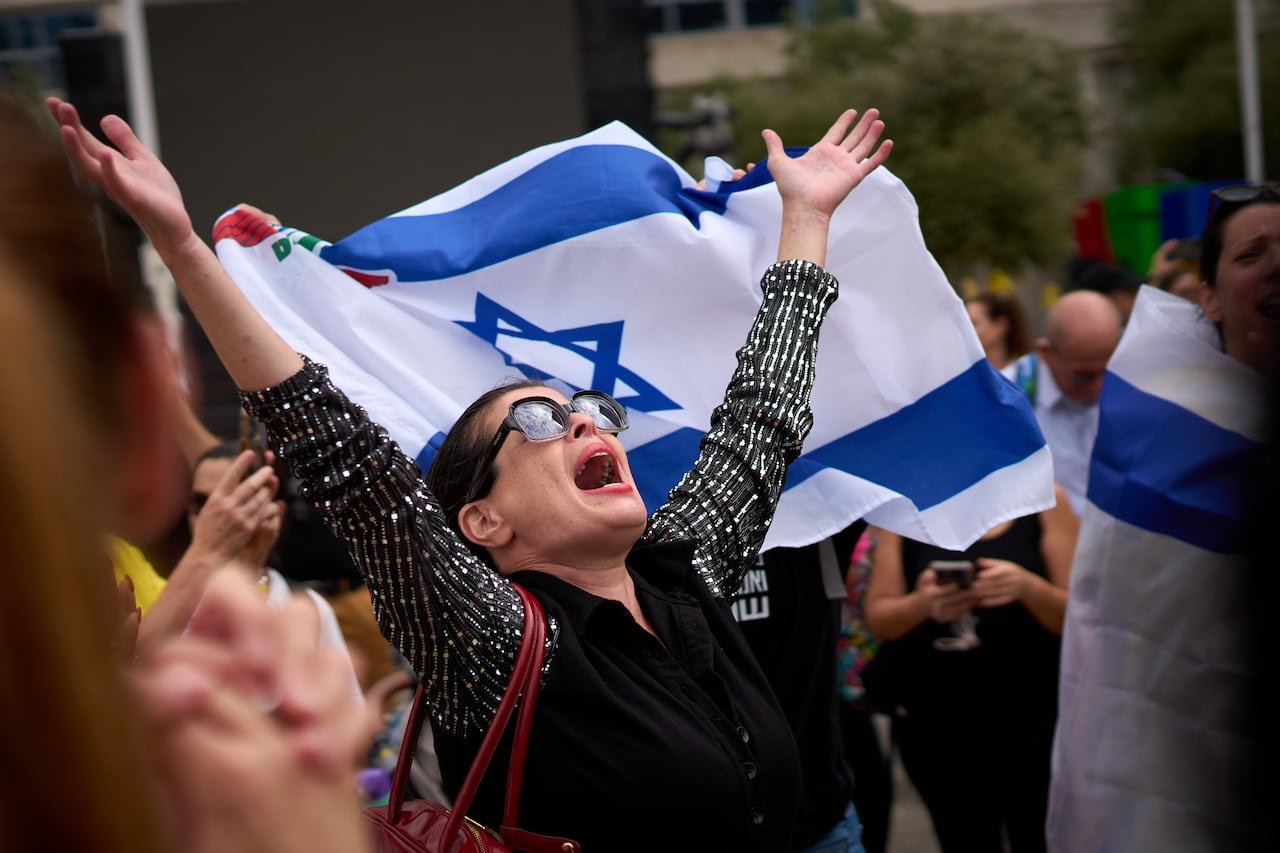 A woman with dark hair in sunglasses extends her arms fully in apparent celebration in front of large flags of Israel.