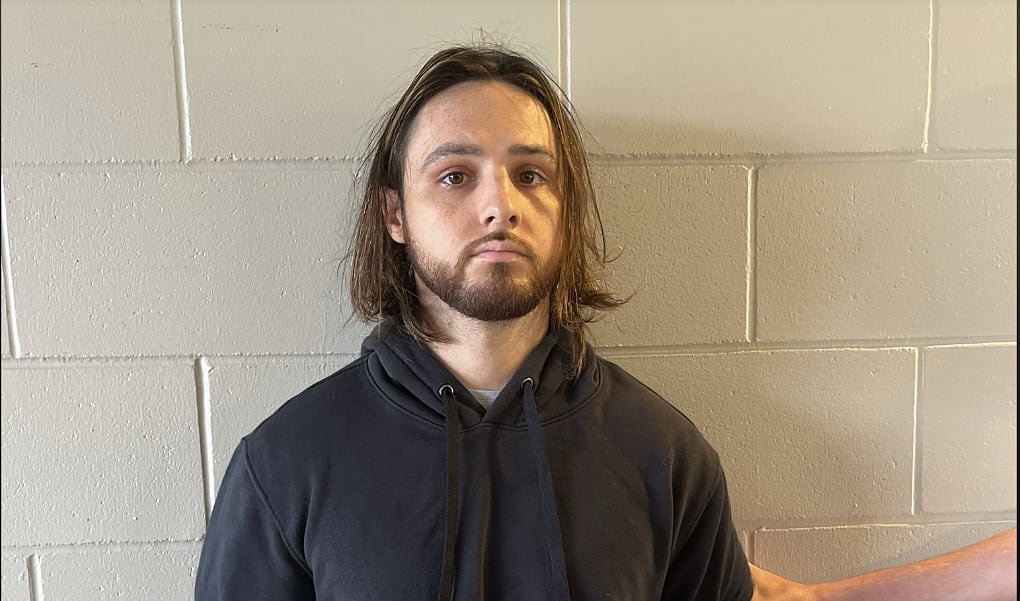 A white man in a black hoodie is photographed in front of a cement wall.
