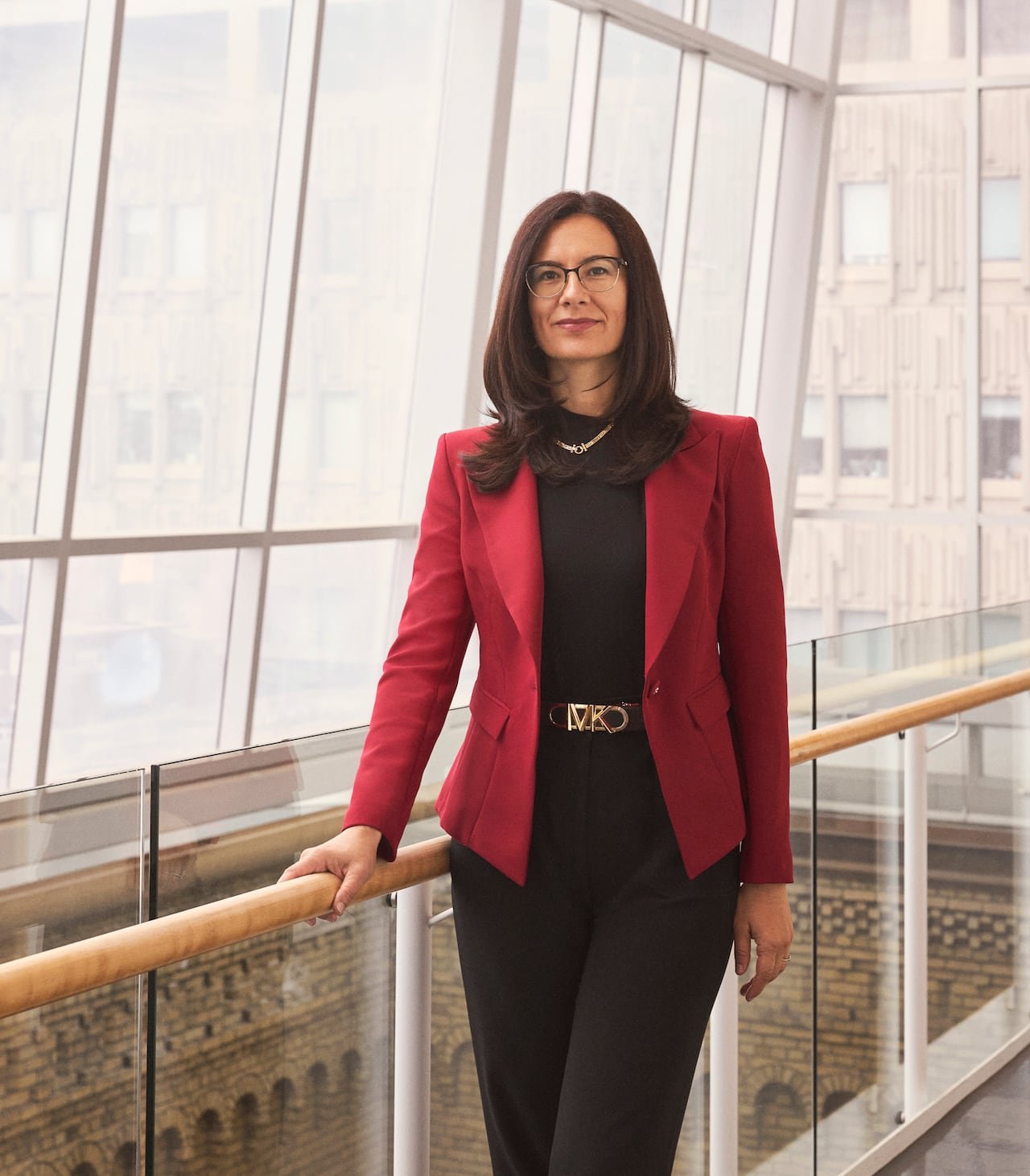 A woman with shoulder length brown hair wearing black with a red blazer and golden chain and belt stands with her right hand on a railing beside large windows.