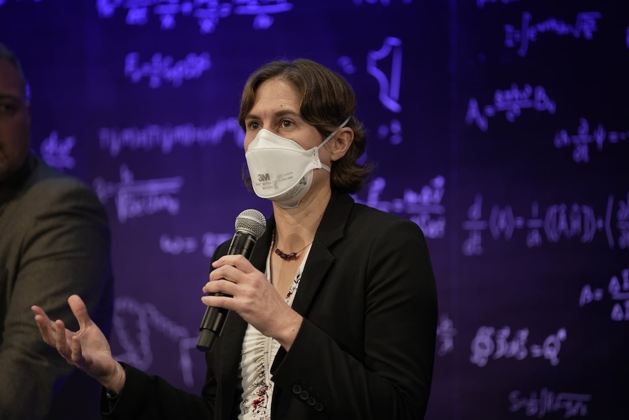 A woman with short brown hair speaks into a microphone on stage.