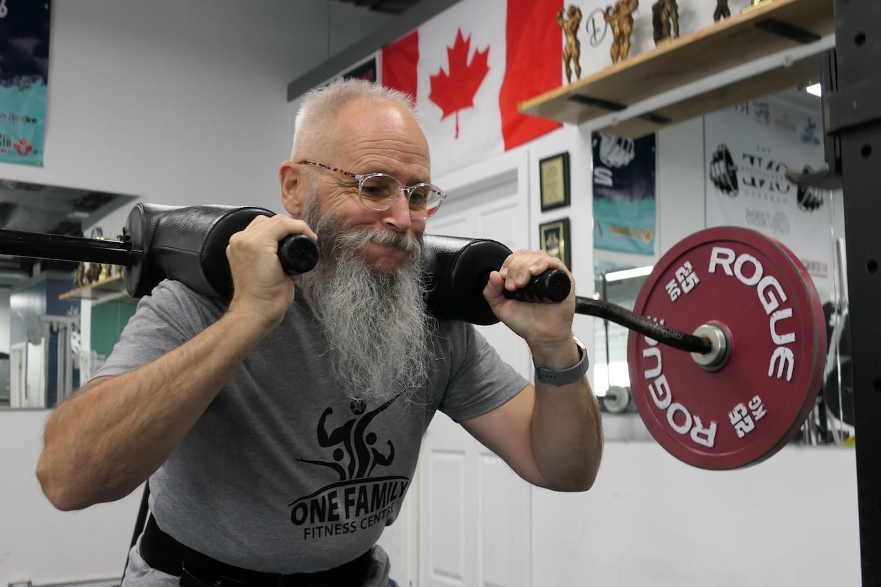 A man in a grey T-shirt, glasses and a big, grey beard squats with weights over his shoulders. His cheeks are puffed.