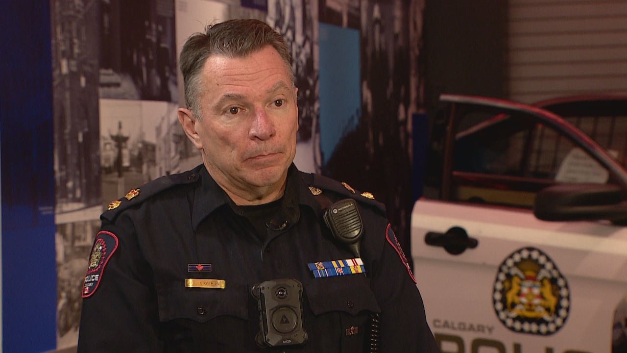 A man with short dark hair that's greying at the sides is wearing  a police uniform sitting in front of a police cruiser with open doors.  