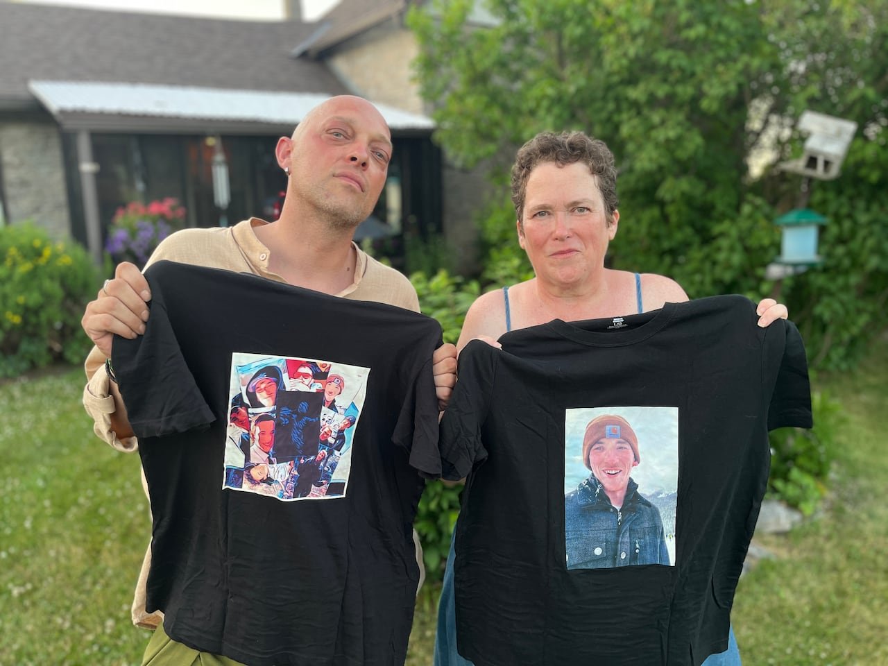 A bald man and a woman with short brown hair stand outside on a sunny summer day holding up tshirts printed with a photos of their son who died at work.