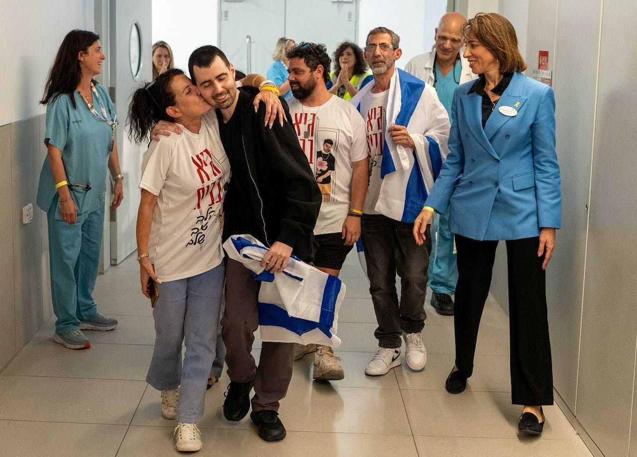 A group wearing matching white T-shirts walks through a hospital with a man wearing a black hoodie. A woman on his right kisses him on the cheek.