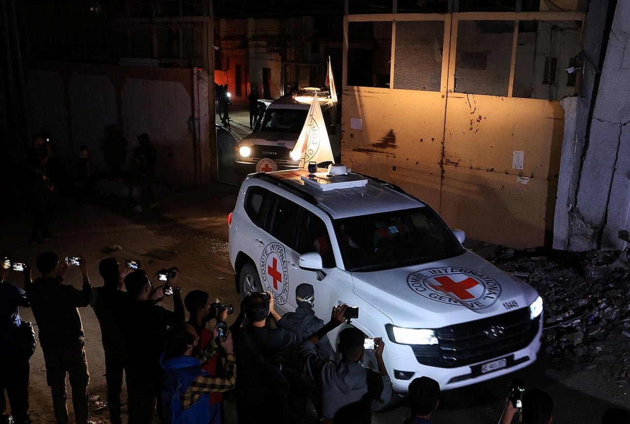 People gather in front of a white Red Cross van at night, taking photos. Another van lines up behind it.