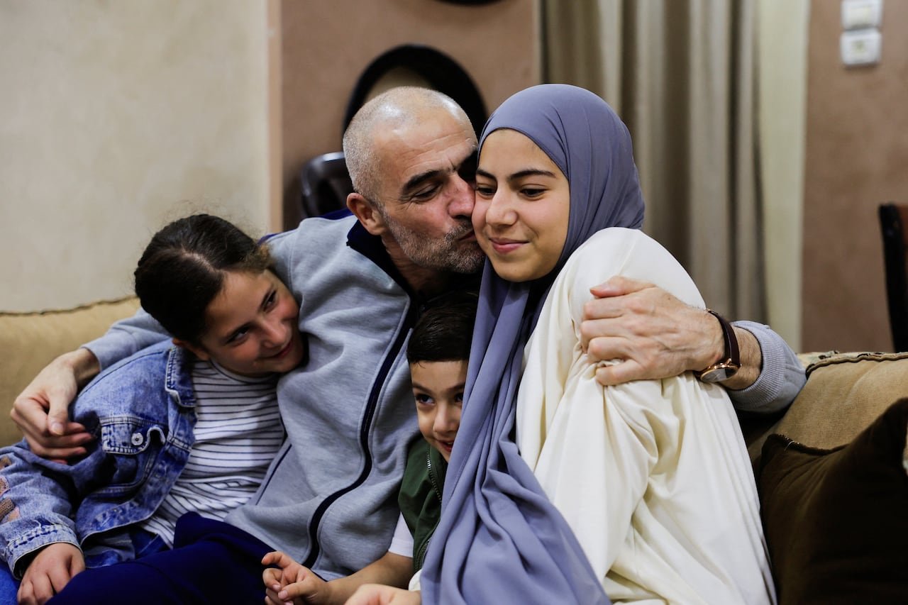 A father in a grey zip-up embraces and kisses his daughter, who is wearing a hijab, while his two younger children lean in for a hug on a couch.