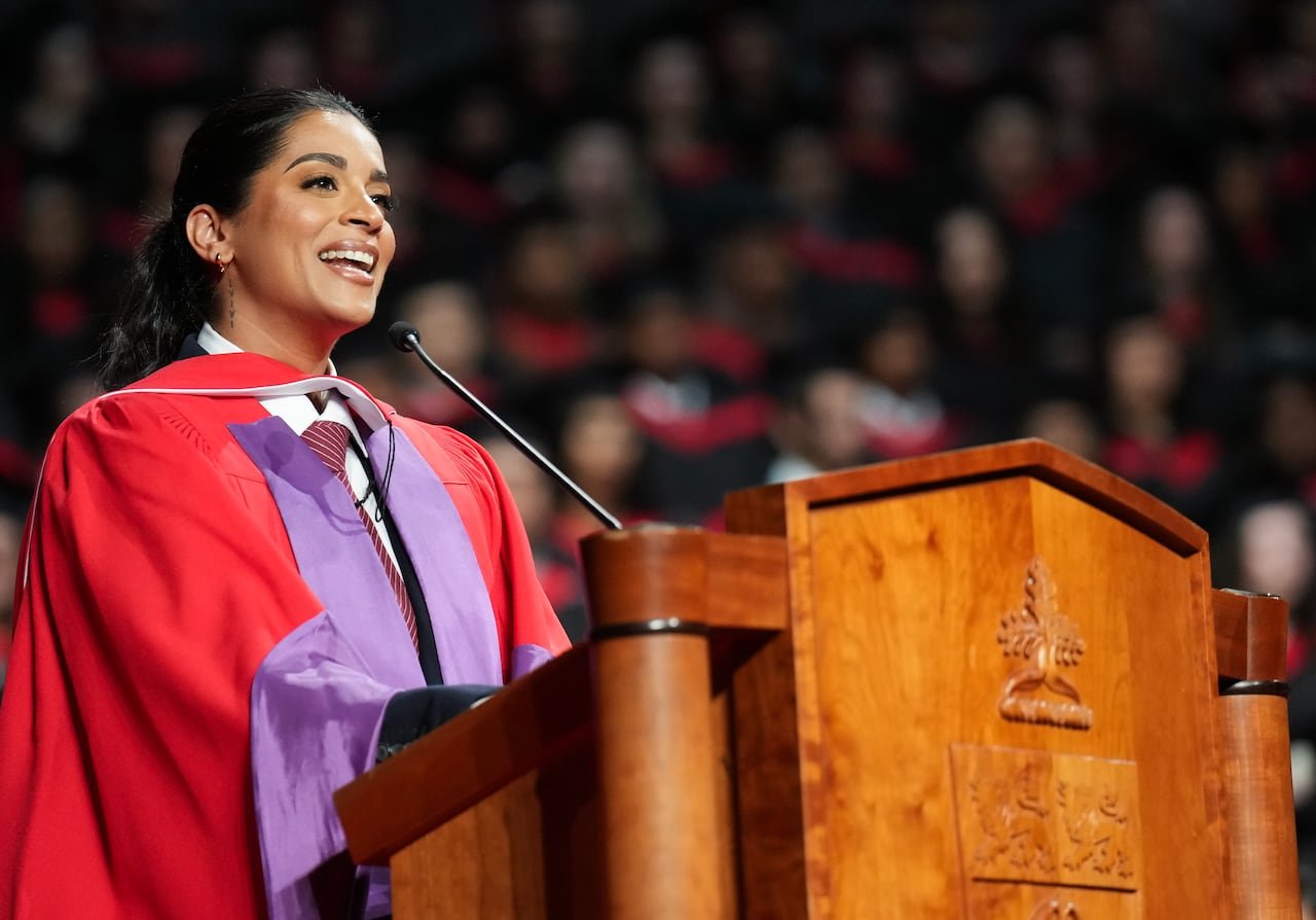 A woman in a red and purple graduation robe speaks into a mic at a wooden podium.