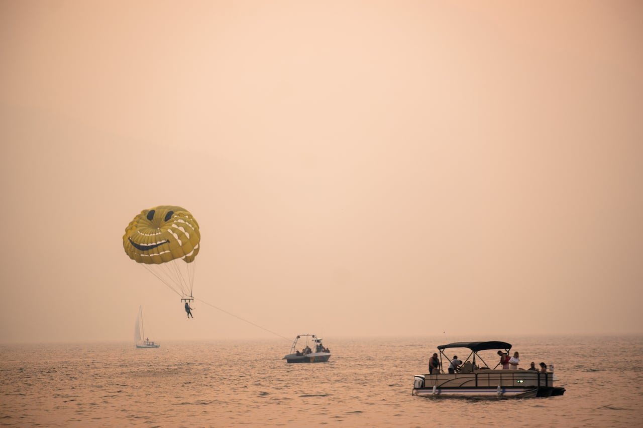 A parasailer with a yellow happy face parachute and three small pleasure boats are shown on a lake. The sky and water are tinted a pinkish orange from wildfire smoke.