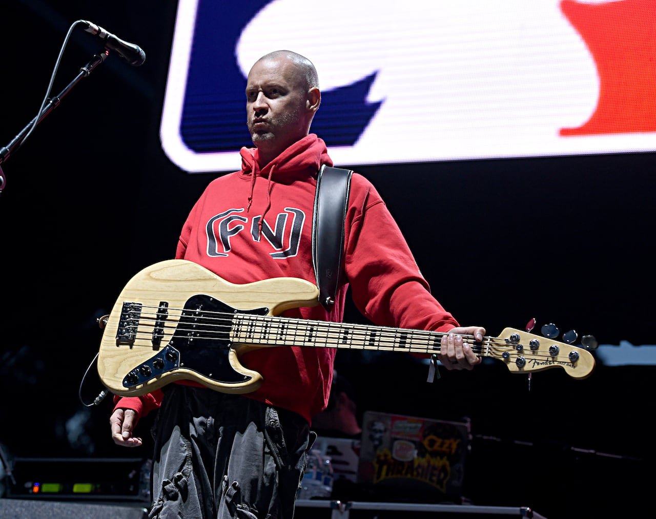 A bald man in a red hoodie stands on stage at night with a bass guitar strapped over his shoulder. 