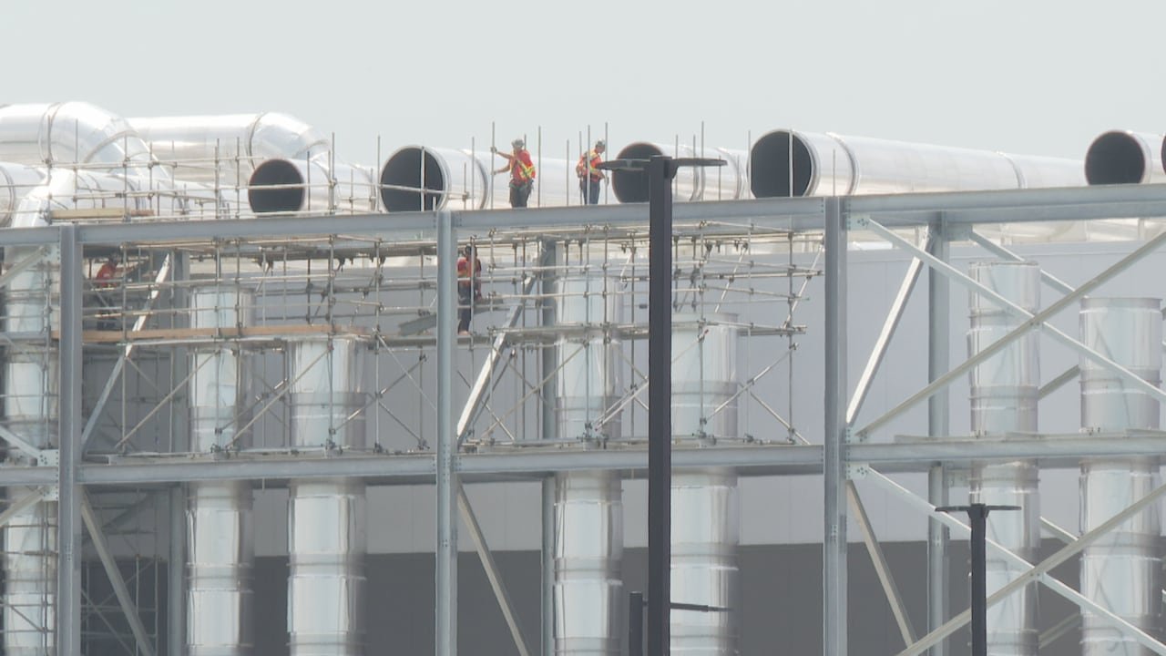 Three workers wearing orange atop a large industrial factory with metal tubing