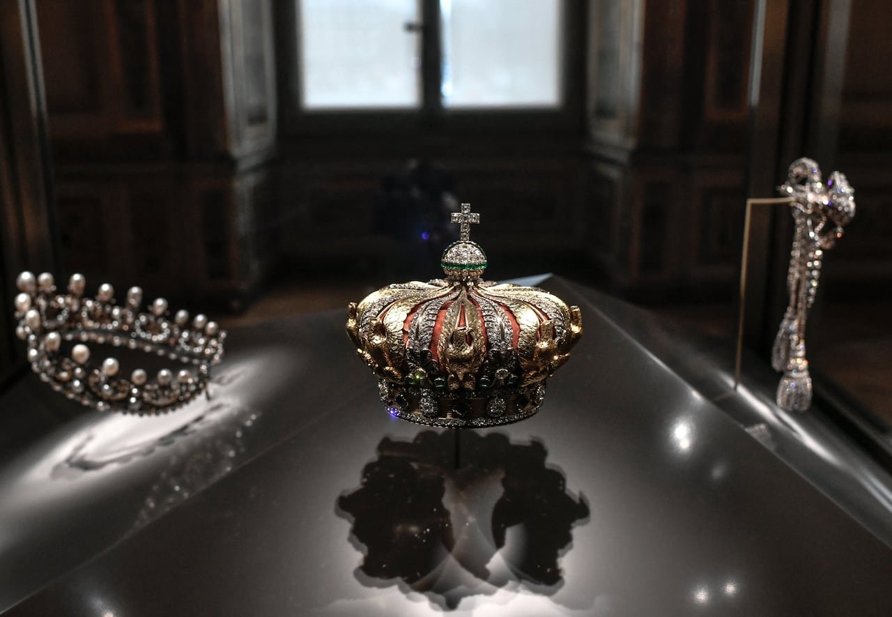 A gold and red crown with a jewel-encrusted orb and cross on top, in a display case.