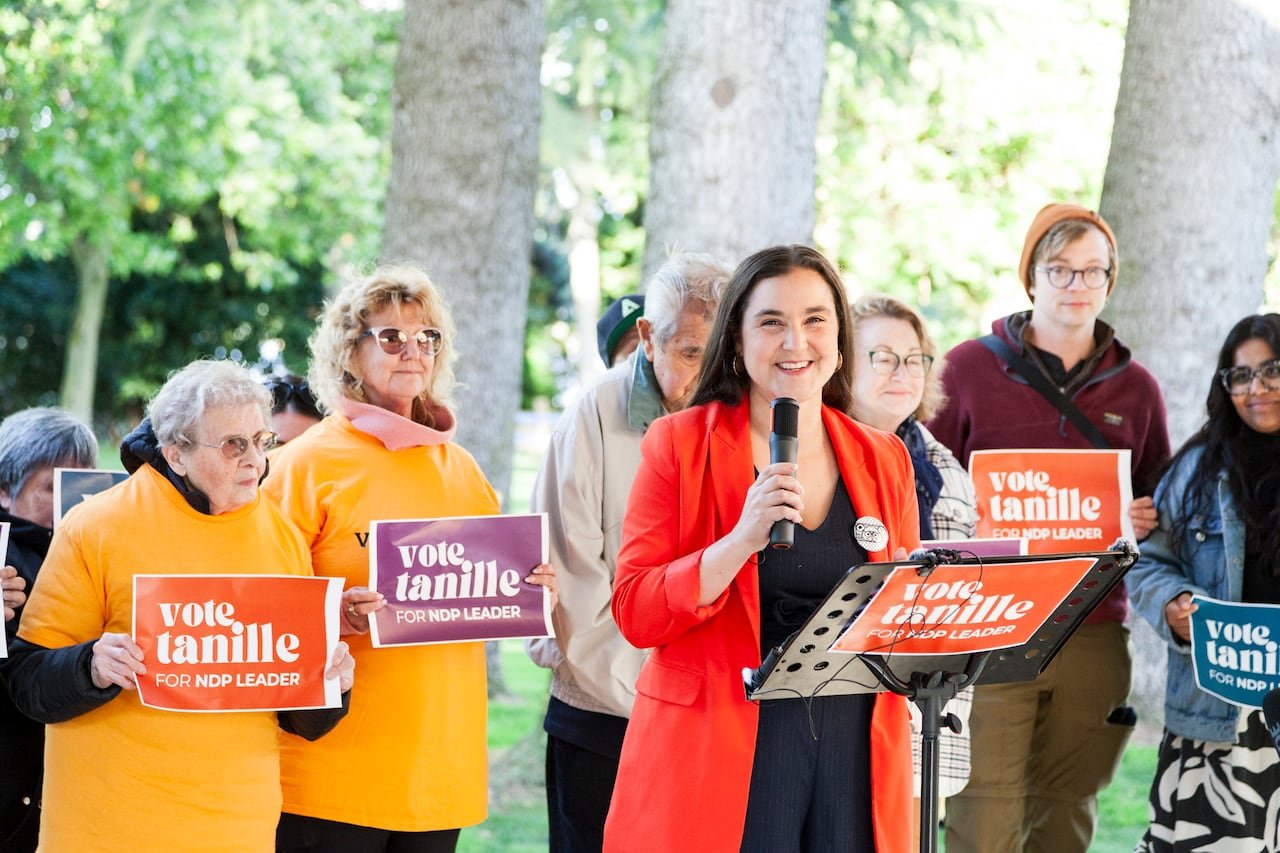 A woman in a orange blazer stands in front of a music stand with folks standing around her.