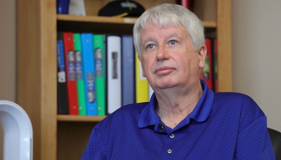 A man in a blue shirt sits in front of a bookcase.