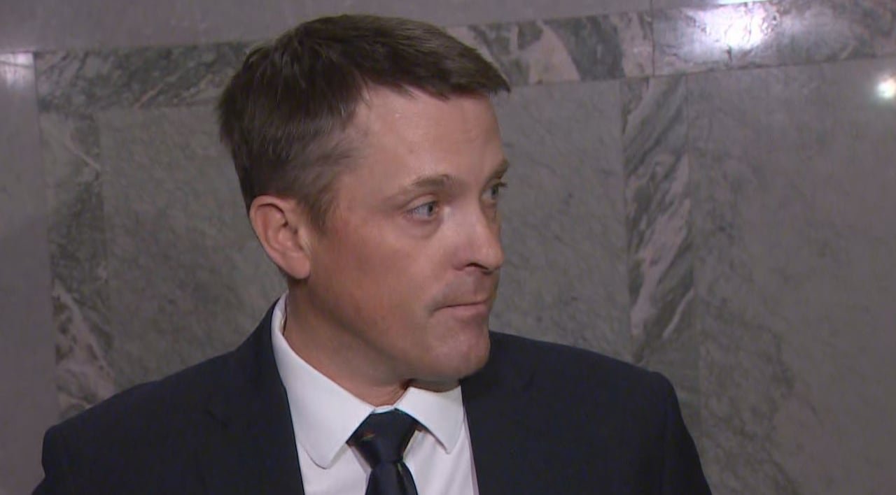 A white man with short dark hair is wearing a dark blue suit and tie, with a white dress shirt. He is standing indoors, in front of a marble wall.