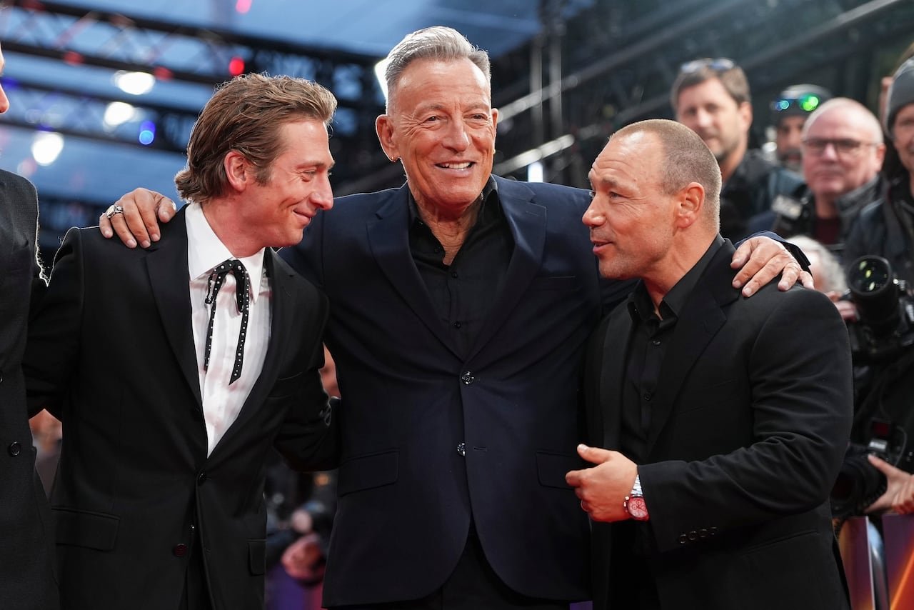 A broadly smiling man in a suit holds his arms over two men beside him. The two men on either side appear to be speaking to one another.