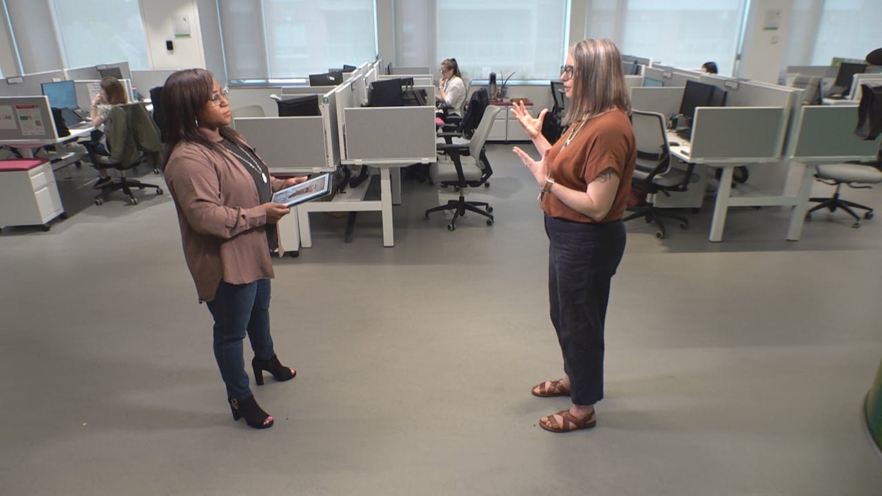 Two women stand in an office space, talking to one another. One is holding a digital tablet.