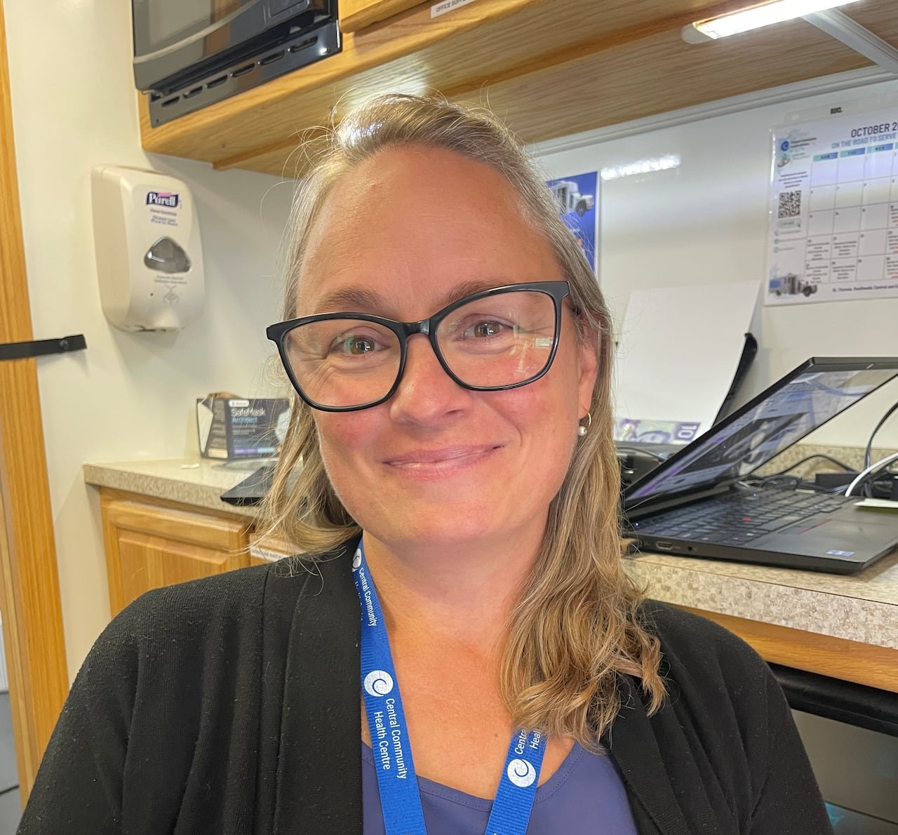 A woman smiles with lips closed, blonde, shoulder length hair and glasses wearing a blue top, lanyard and black sweater. 