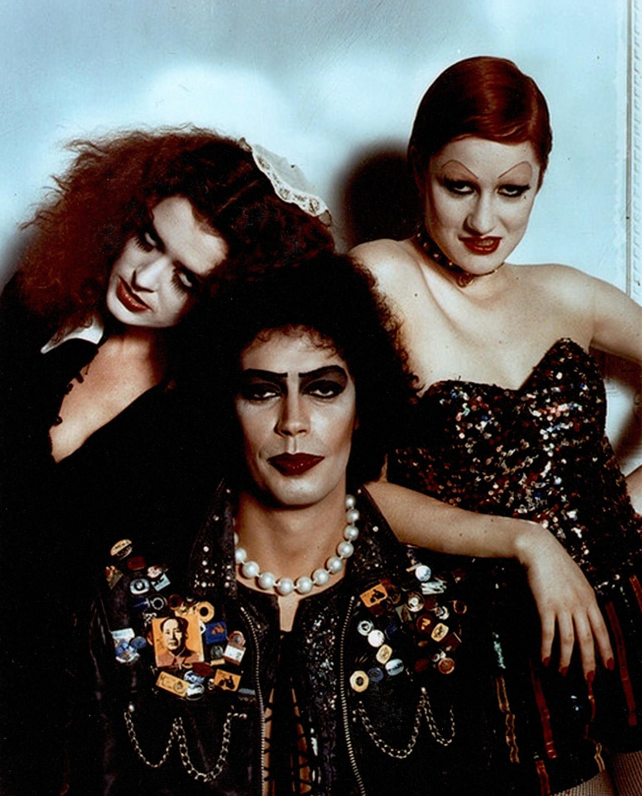 Three people with sultry facial expressions and unique, flashy costumes stare into the camera against a blue backdrop.