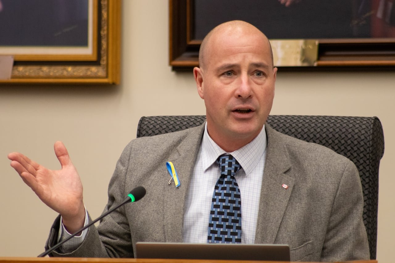 A person wearing a suit is seen sitting behind a desk speaking into a microphone.