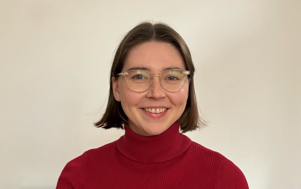 A person with short brown hair, transparent glasses and a red turtleneck is seen smiling.