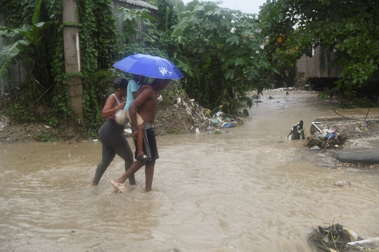 A man carries a child on his back and a woman walks behind as they all crowd beneath an umbrella and walk in ankle deep water.