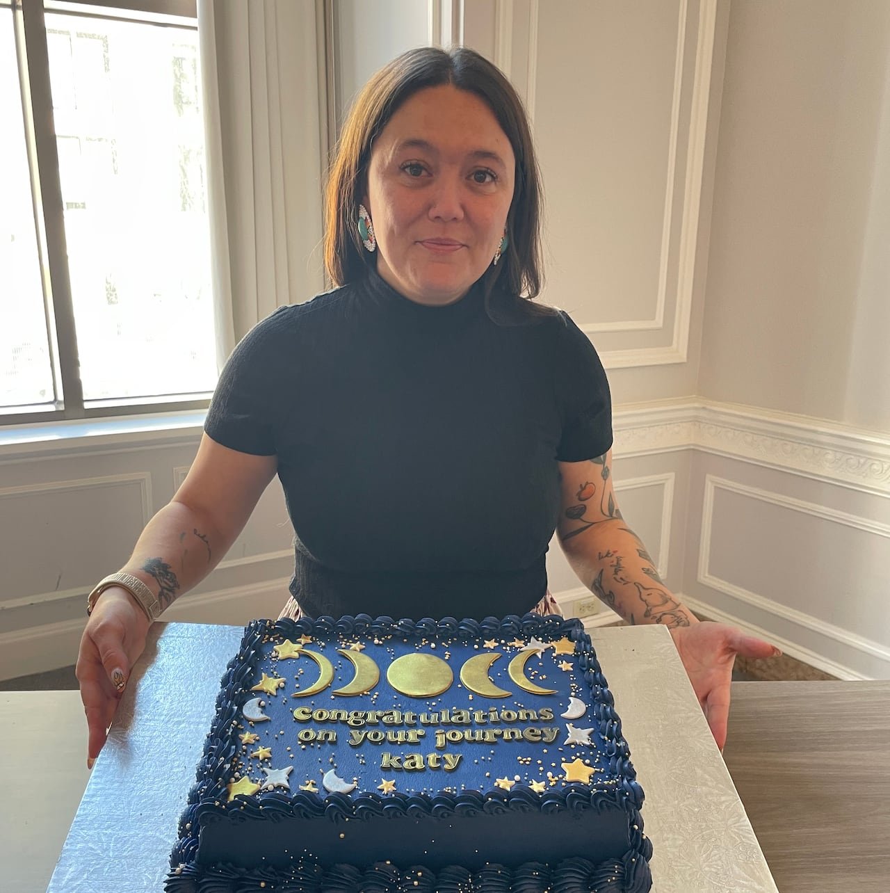 A woman wearing a ribbon skirt smiles at the camera while standing in front of a blue cake that reads "congratulations on your journey Katy."