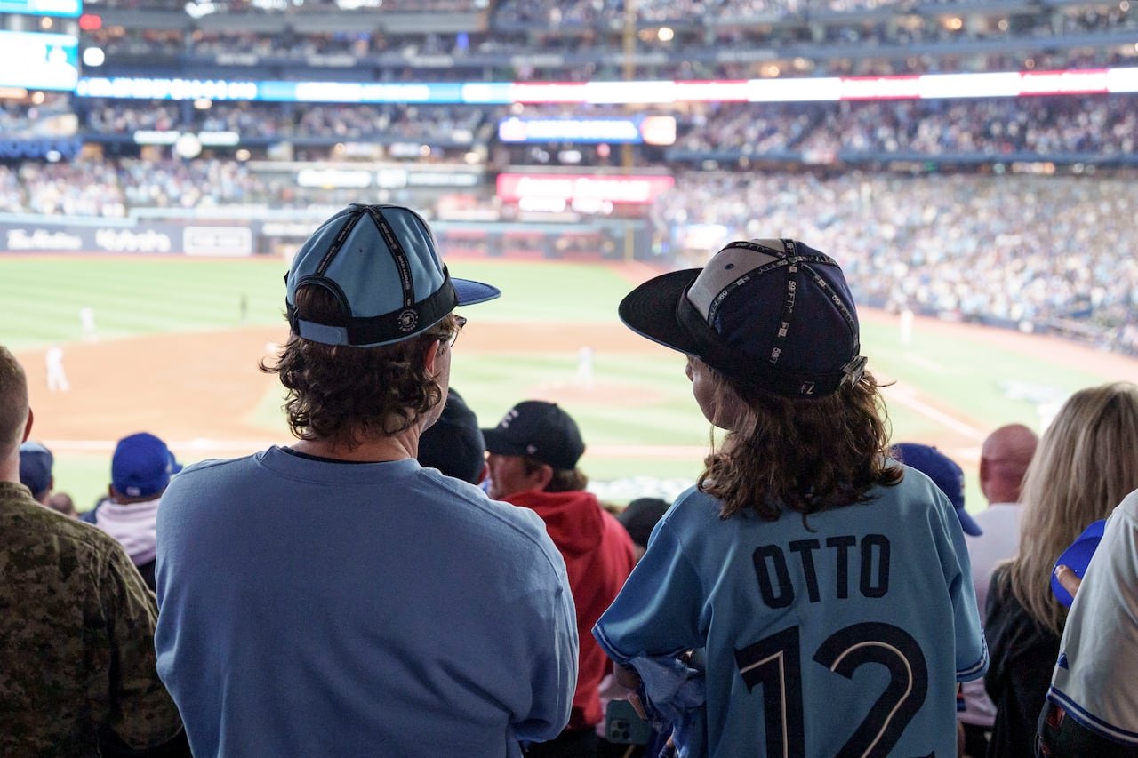 Young girl and her dad sit at Rogers Centre with Blue Jays jerseys on and inside-out Blue Jays caps
