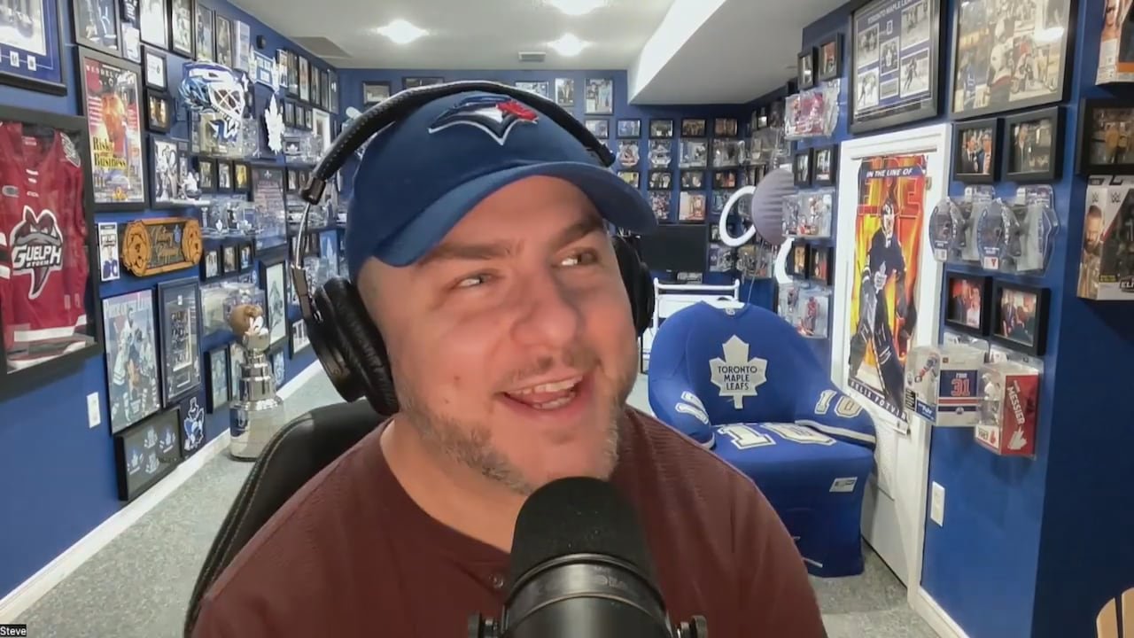 A man in headphones and a Toronto Blue Jays baseball cap sits in a room full of sports memorabilia.