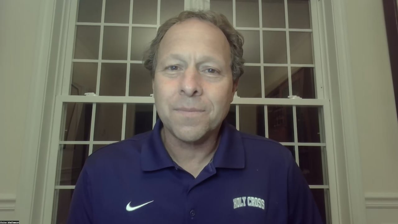 A man in a blue polo shirt labelled 'Holy Cross' sits in front of a window facing a webcam.