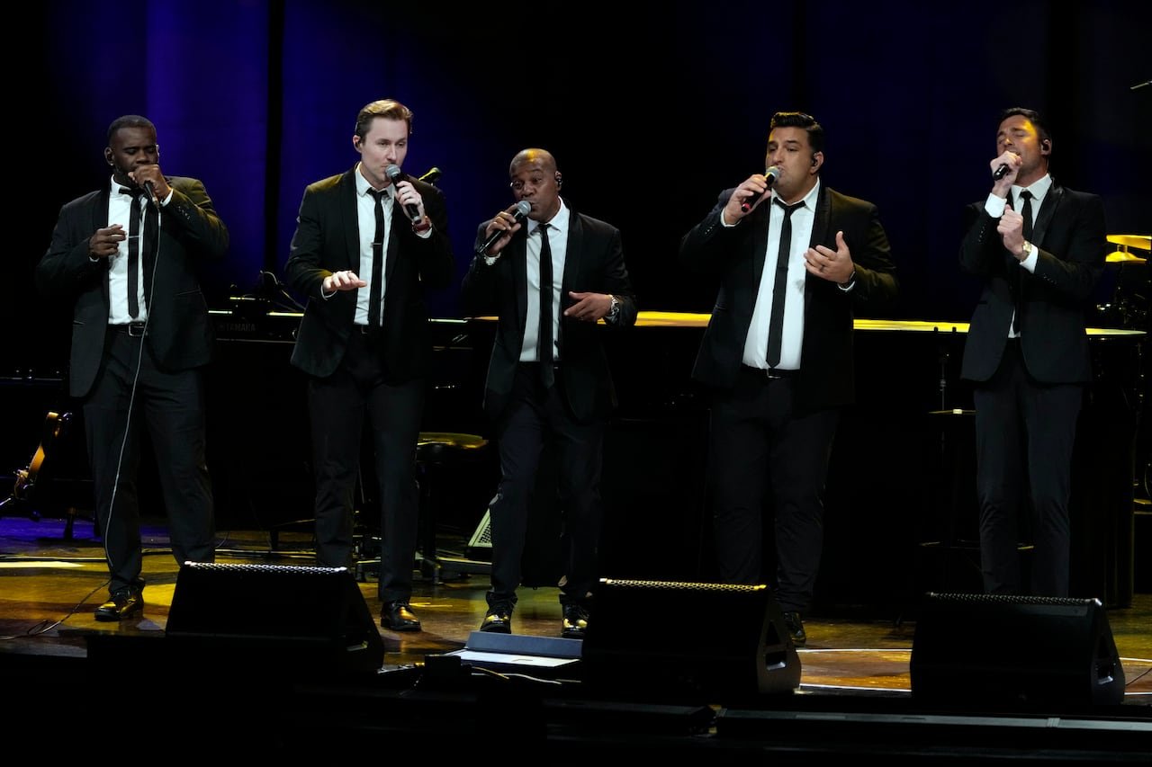 Five men in suits sing into microphones on a stage.