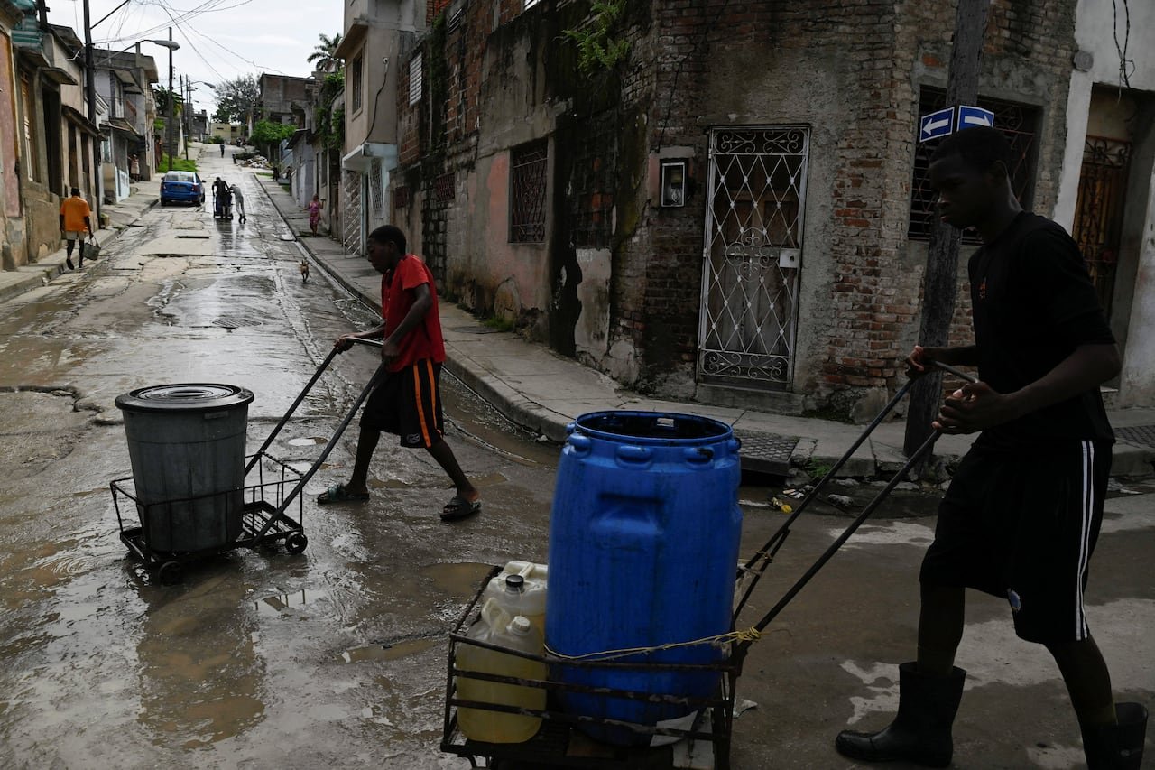 Two dark-complected men wearing shorts wheel barrels through narrow city streets.