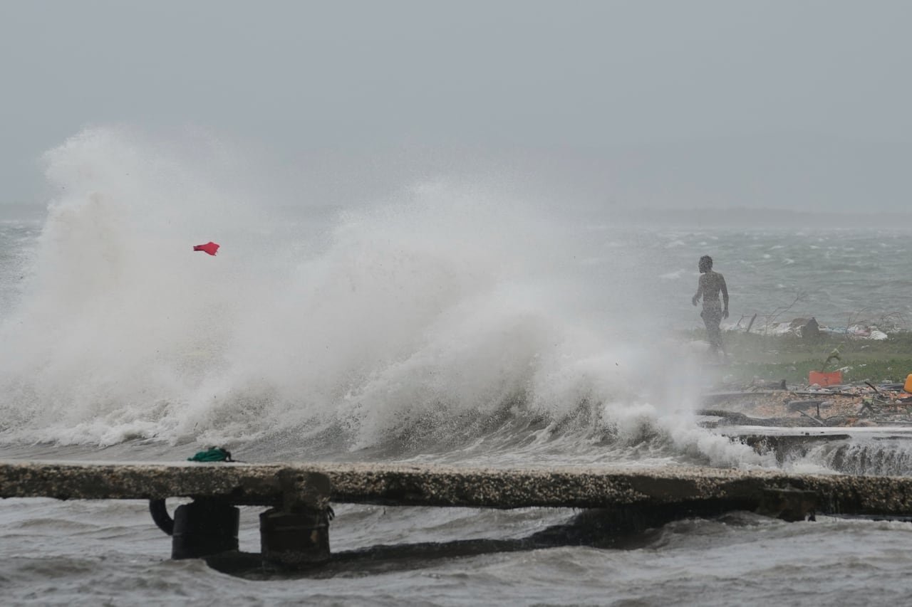 A large wave is seen washing ashore on a cloudy day. In the distance, a dark complected, shirtless man is shown.