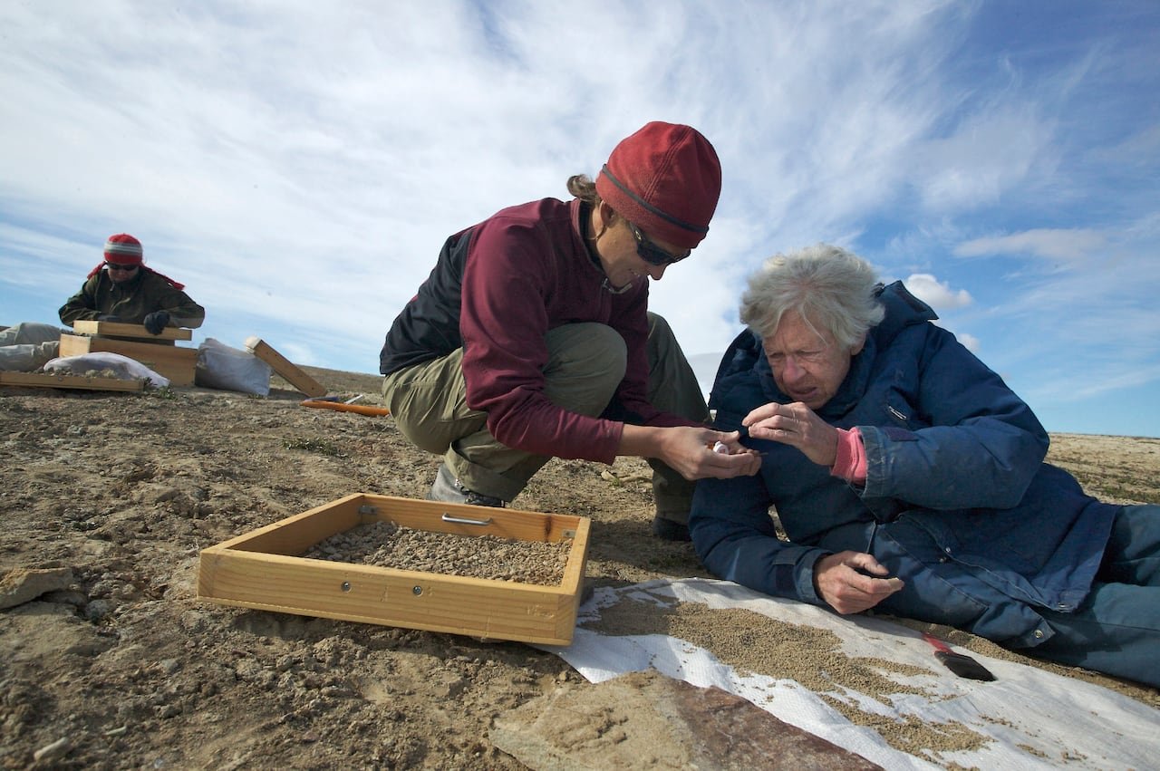 Two women on gravelly ground with a rectangular screen beside them