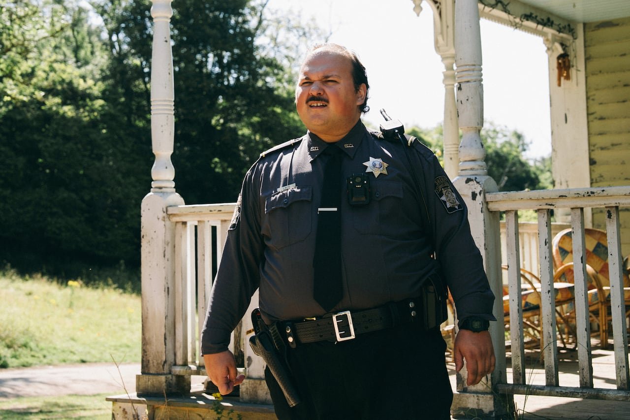 A man wearin ga police uniform stands outside a weathered porch.